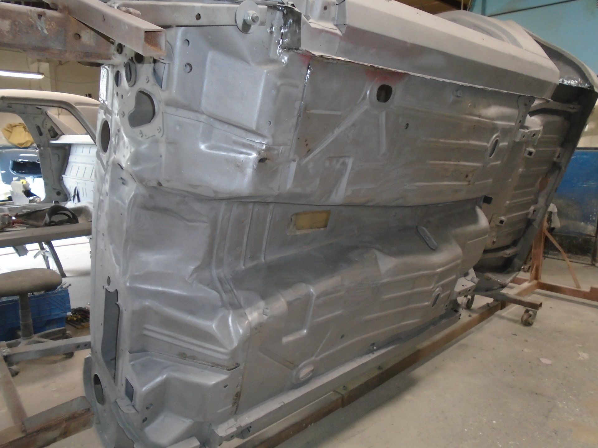 Underside of a car chassis, primed silver, elevated on a support, inside a workshop.
