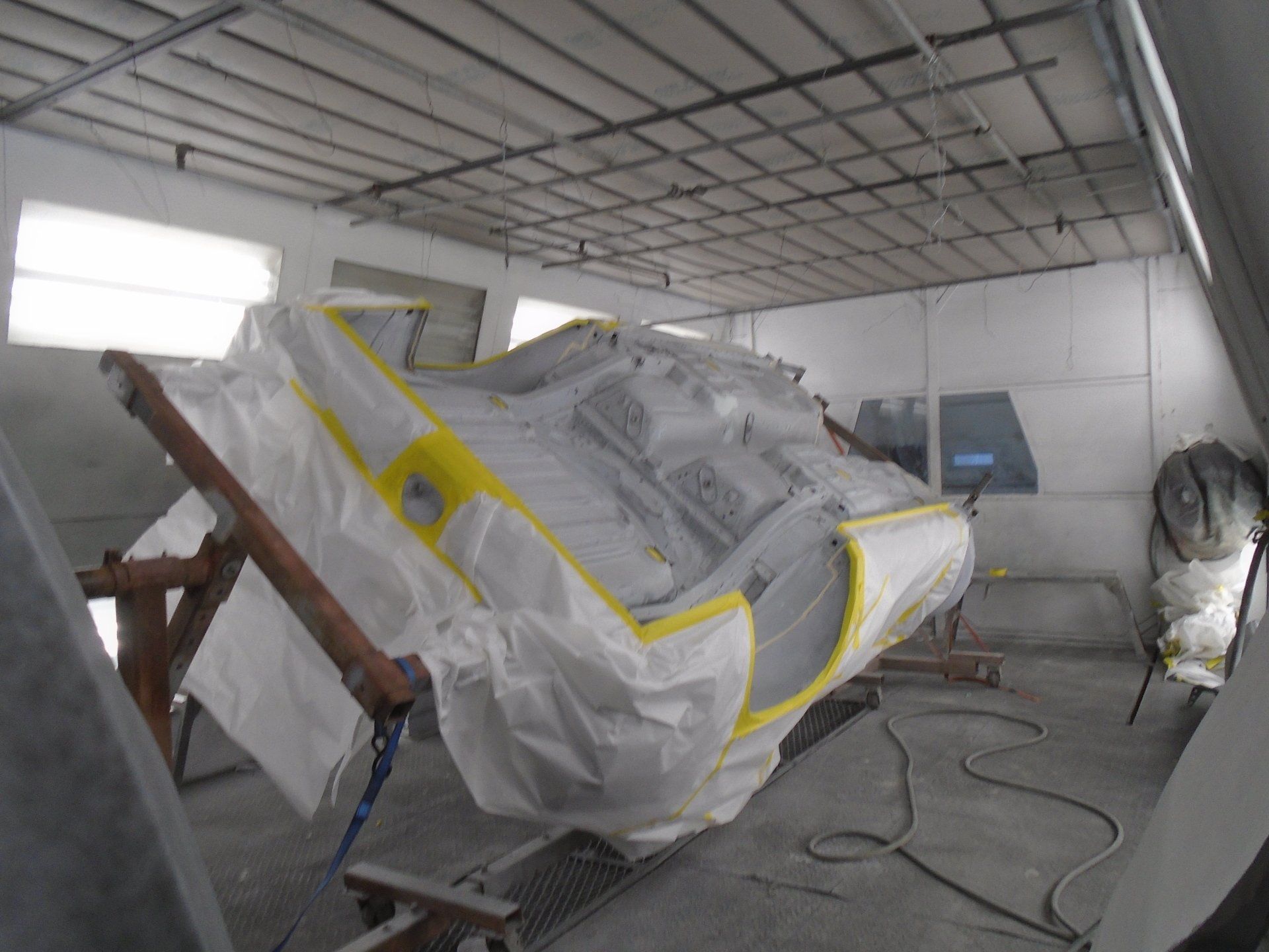 A car's body, covered in white masking paper and yellow tape, inside a spray paint booth.