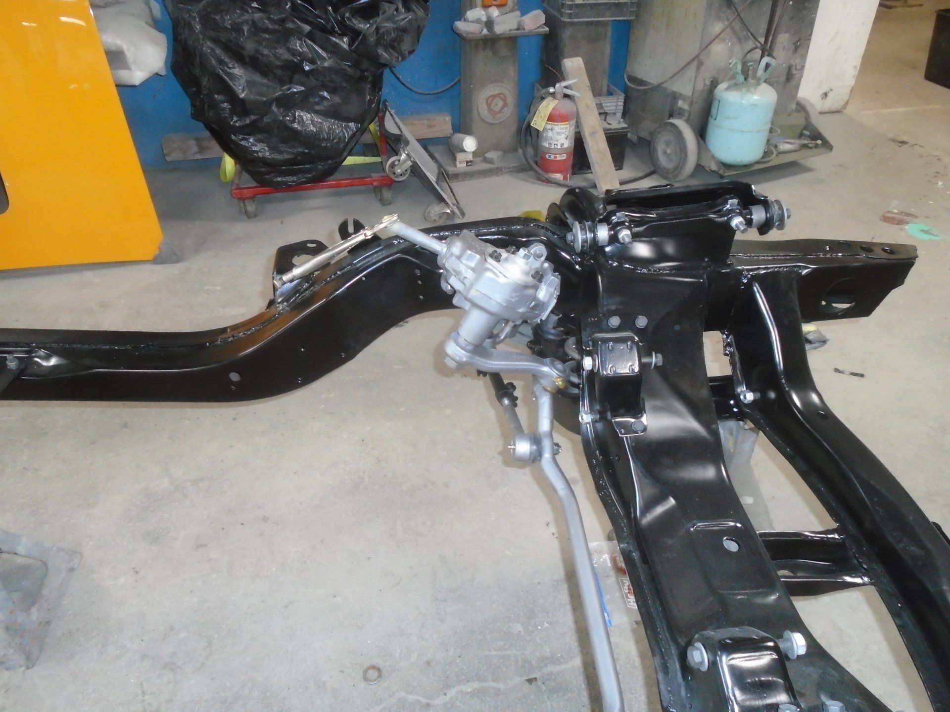 Black car frame with steering mechanism. Yellow panel, various tools and equipment in background.
