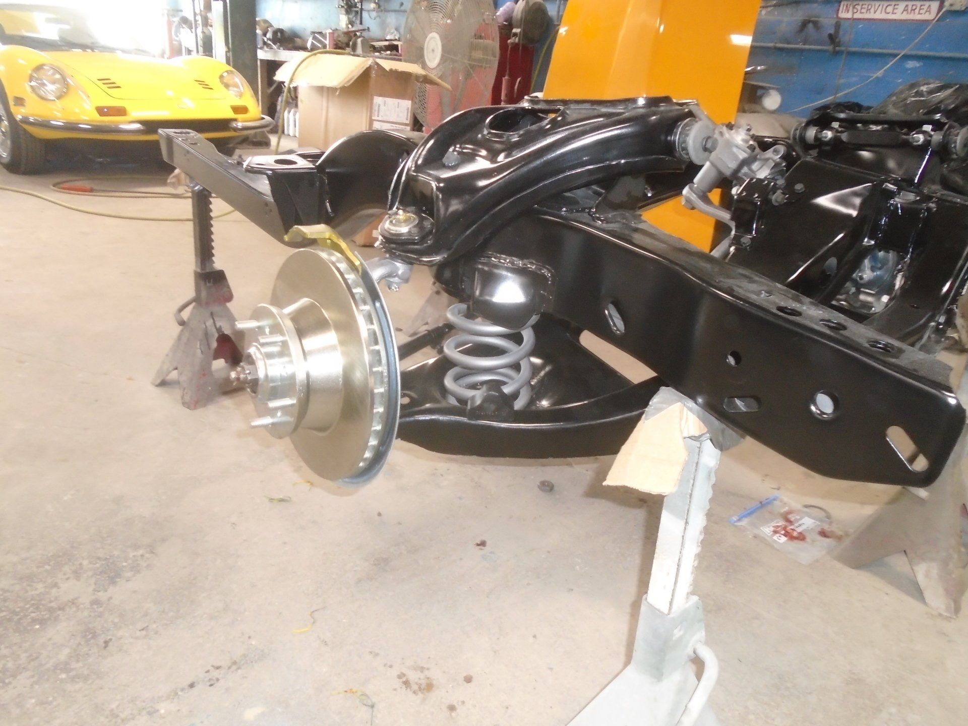 Black car frame on jack stands; front suspension, rotor, and coil spring visible. Yellow car in background.