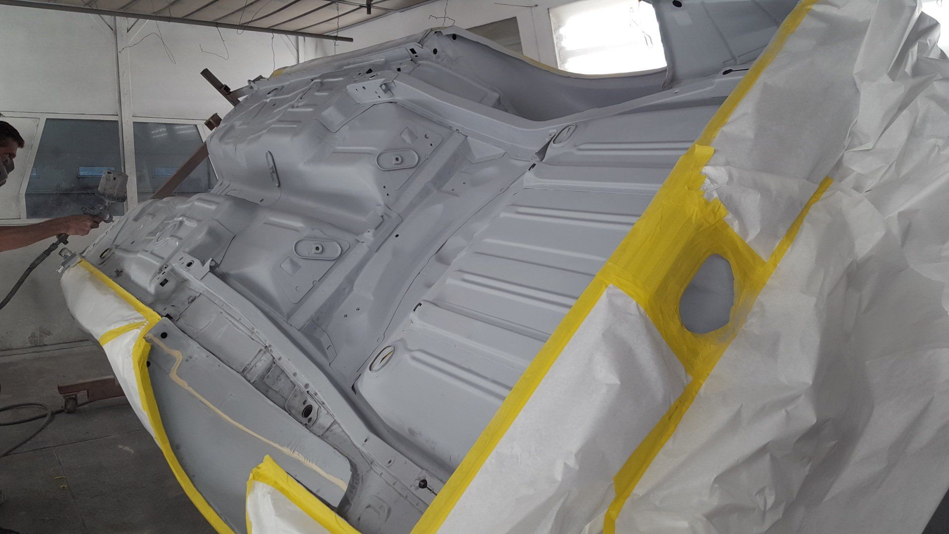Person spraying paint on the underside of a car in a workshop. Car is masked off with yellow tape.