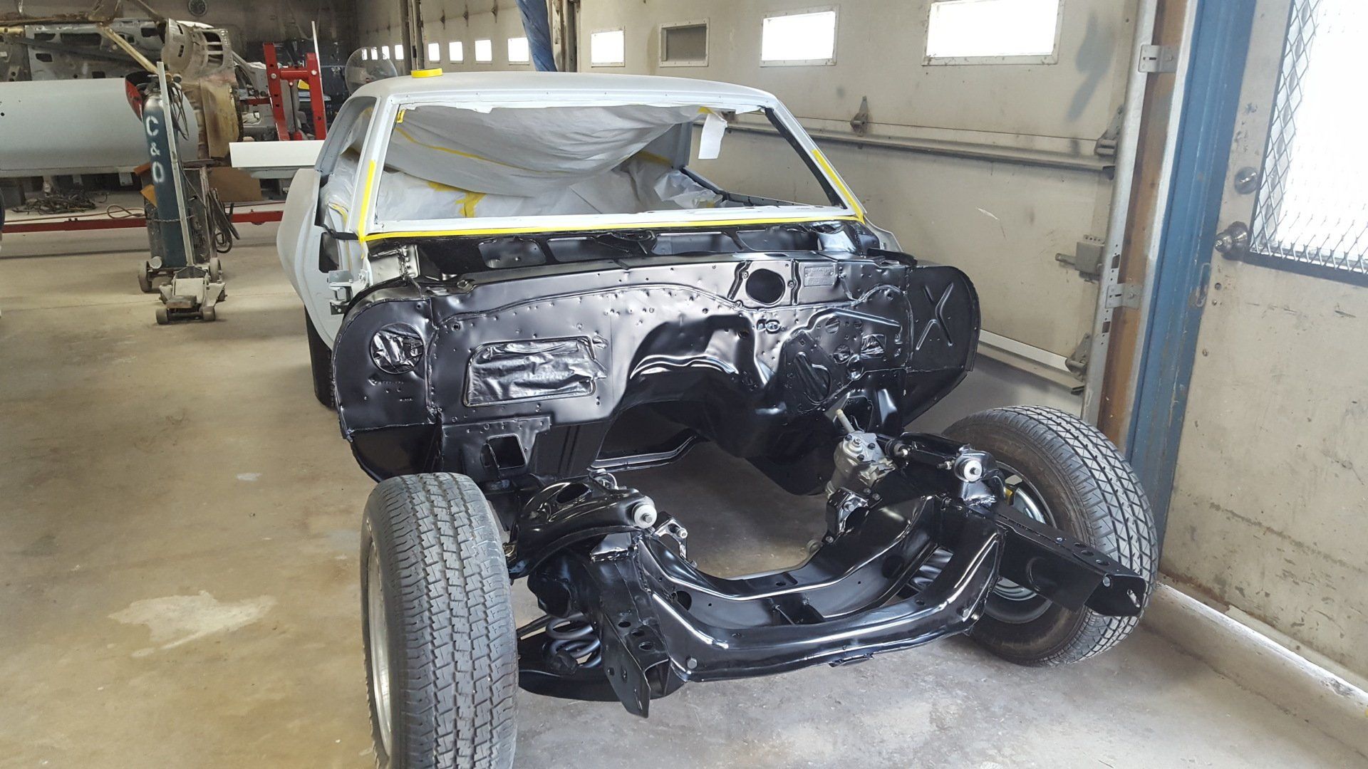 Black-painted car chassis, frame, and body in a workshop. Parts masked for painting.