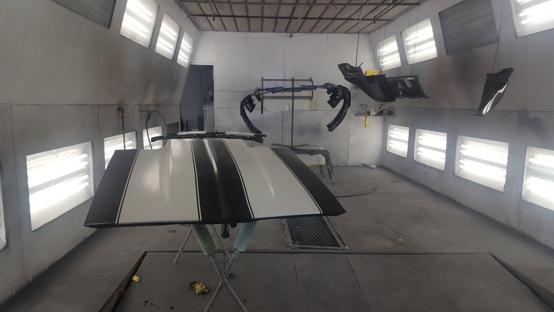 Car parts, including hood, bumper, and panels, being painted black and white in a spray booth.