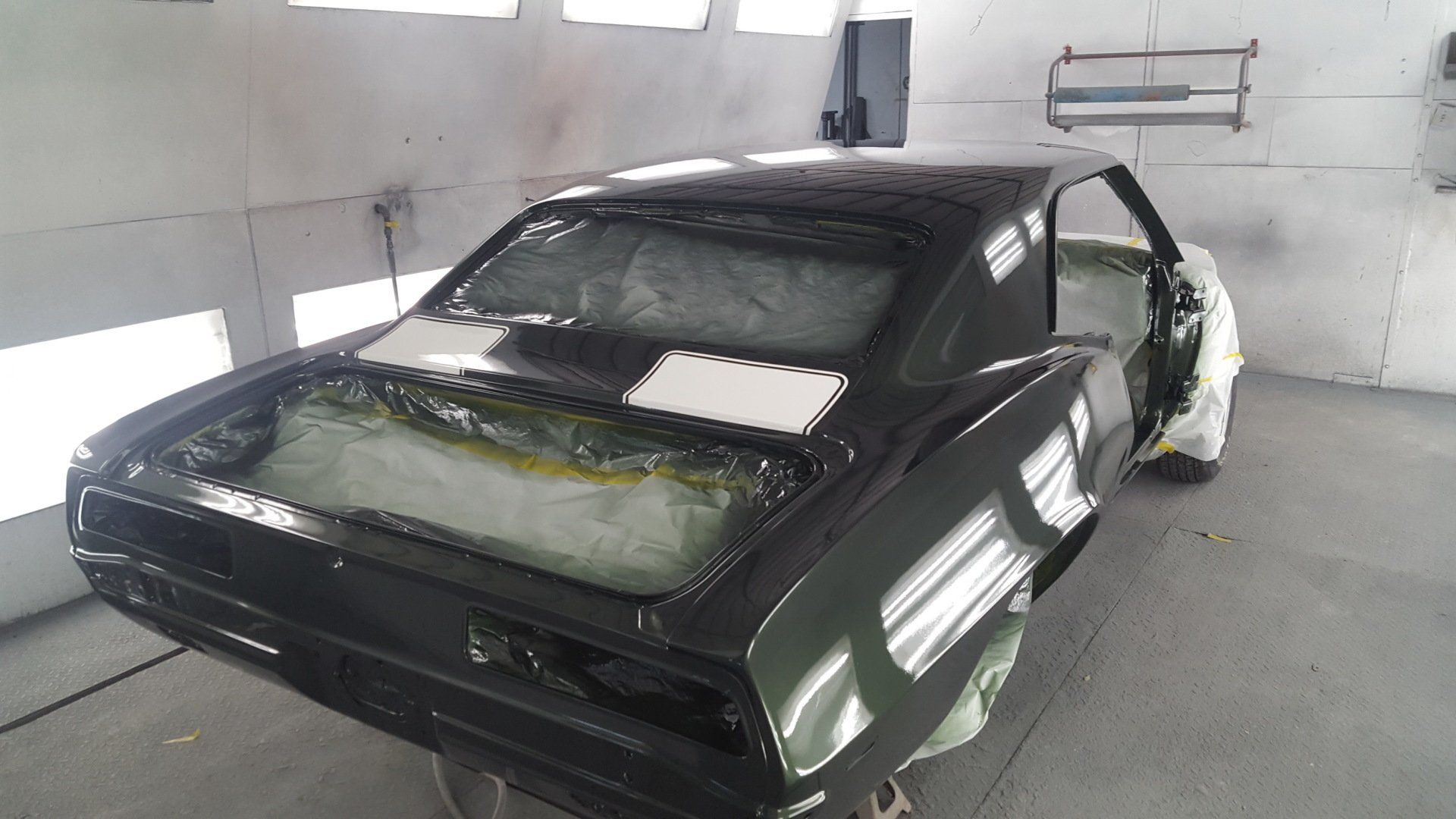Black classic car in a paint booth being restored, with masked areas and shiny finish.