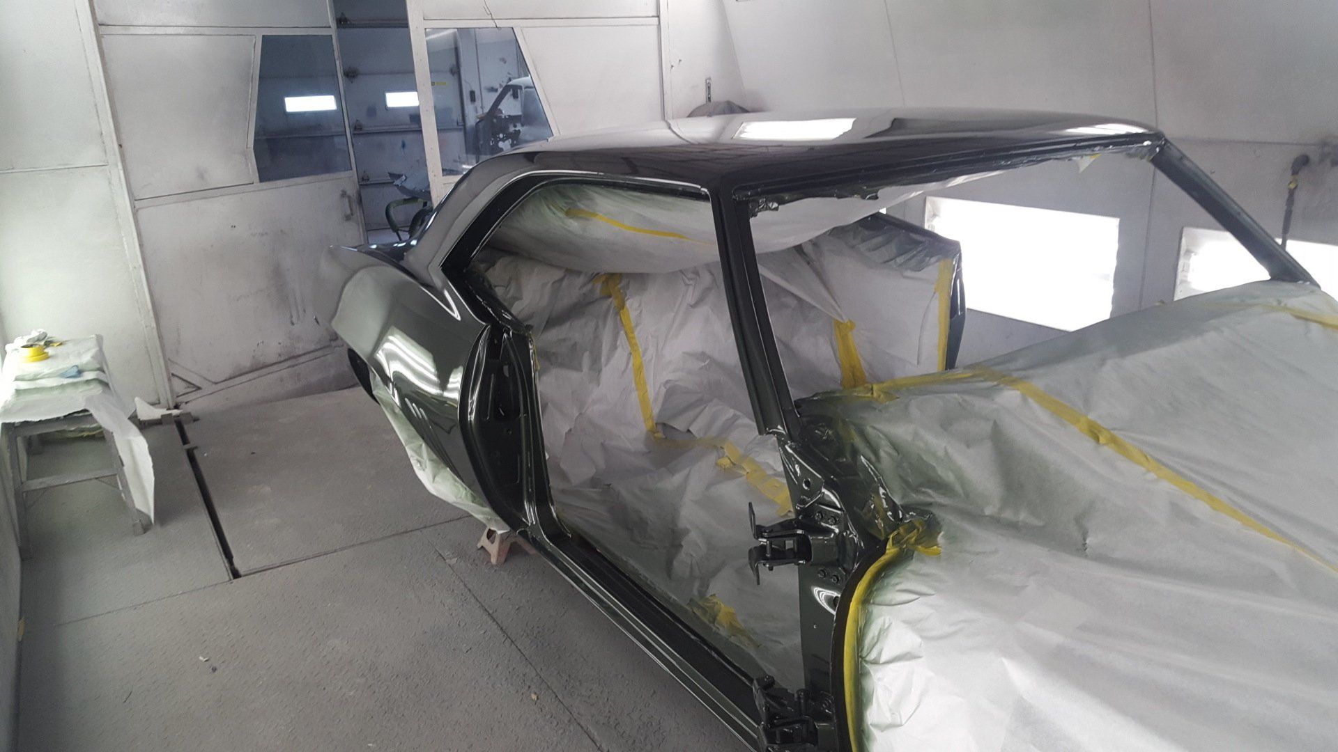 Classic car in a paint booth, partially masked, with new dark green paint.