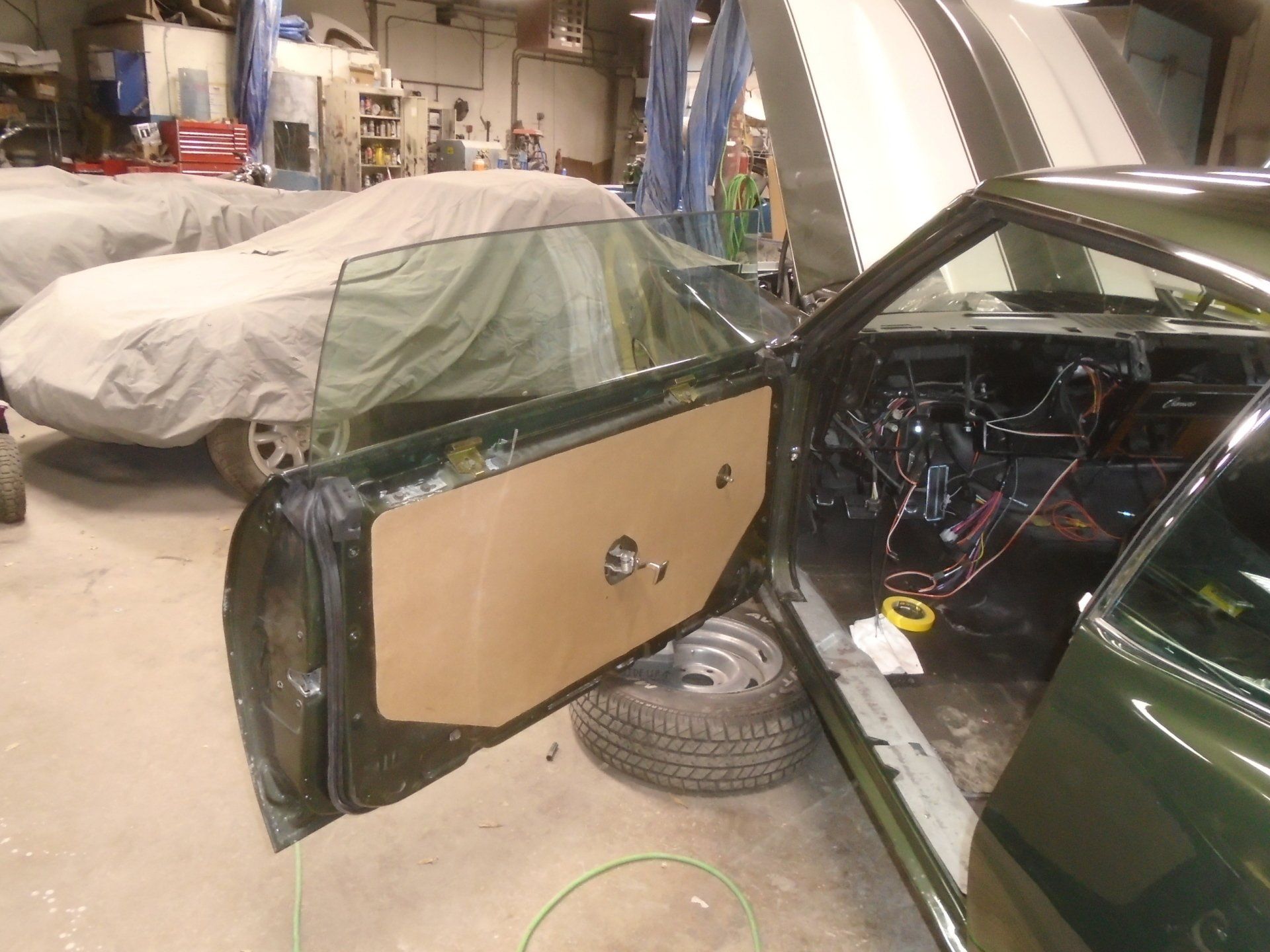 Green car door open, interior exposed. Door panel installed. Another car covered in background.