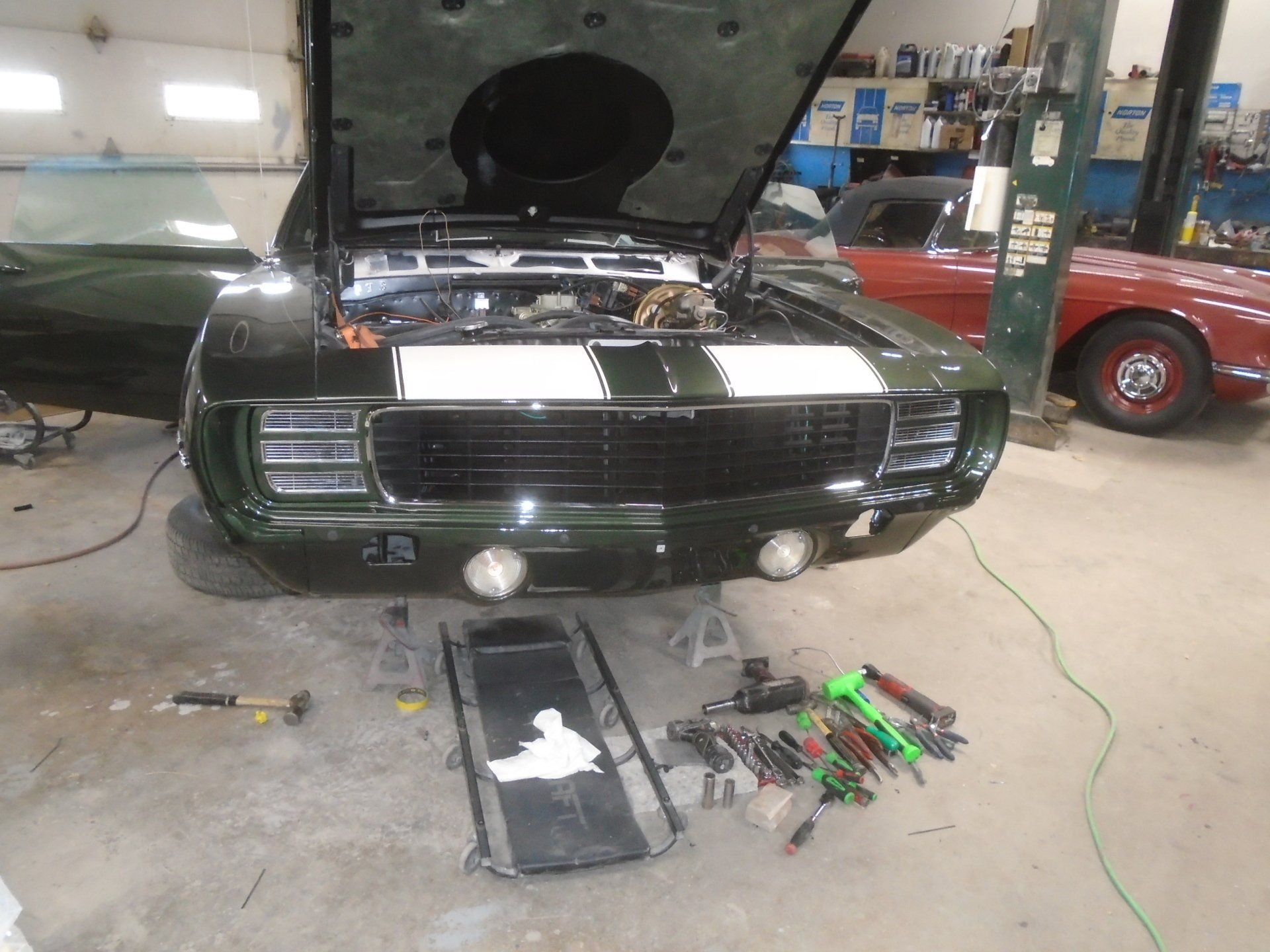 Green classic car with hood open in a garage, tools and parts visible.