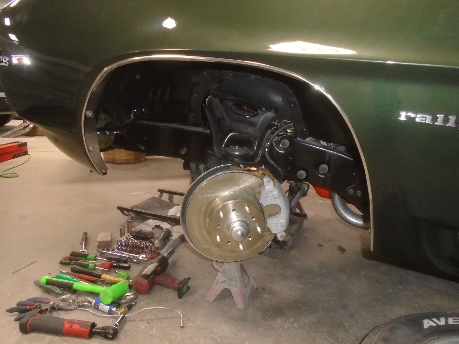 Green classic car with front wheel removed, revealing suspension components and brake rotor. Tools are scattered nearby.