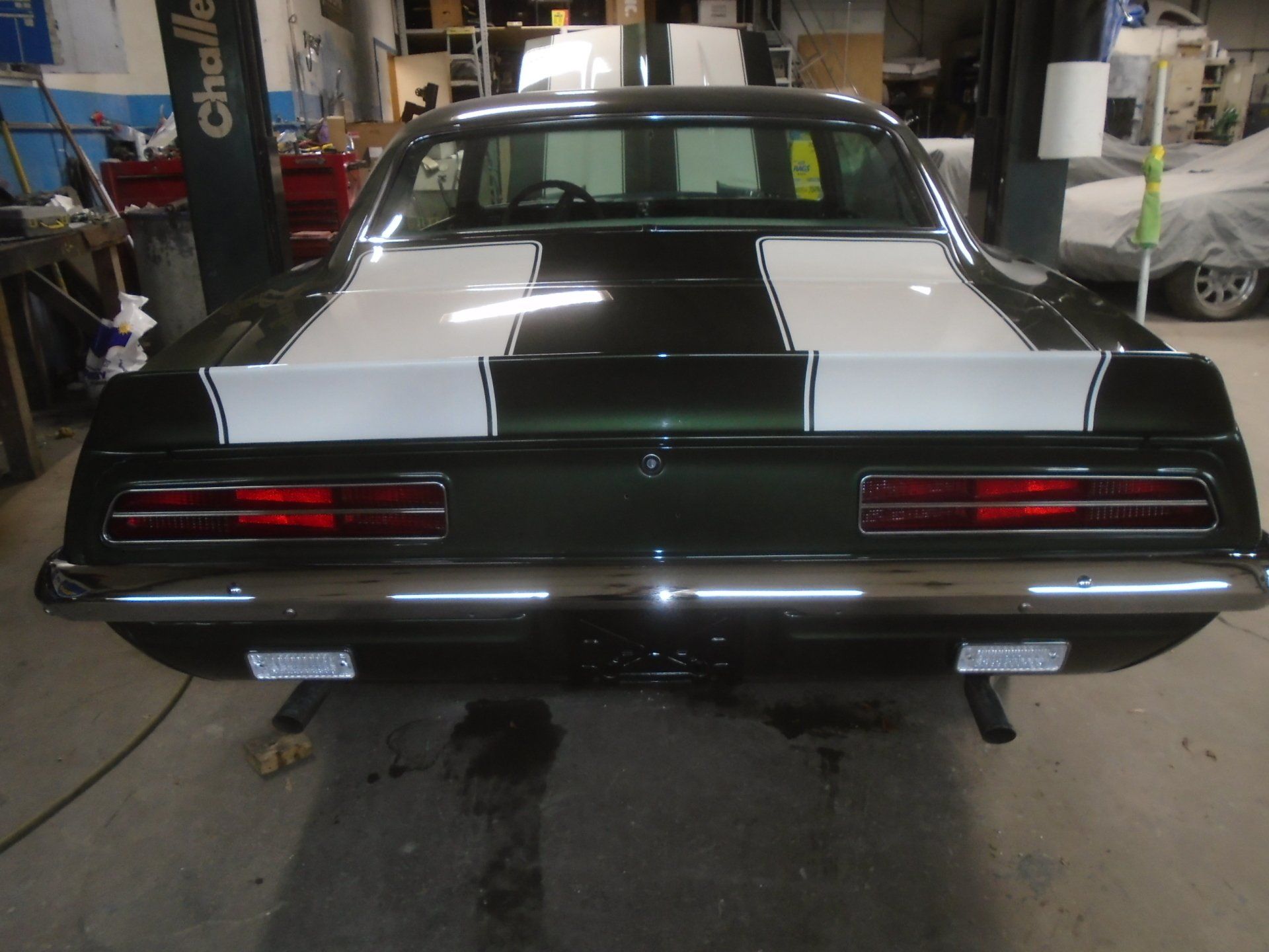 Rear view of a dark green classic car with white racing stripes, in a garage.