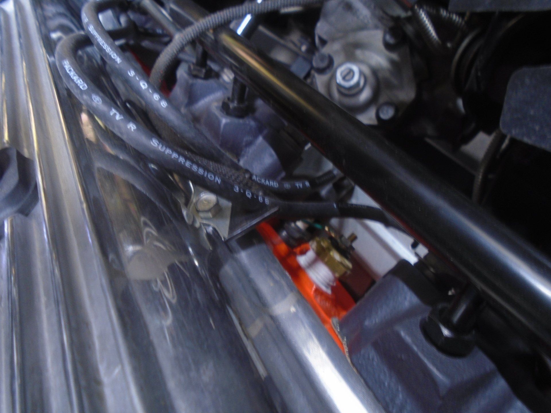 Engine bay, metallic tubing, black hoses, orange block, and black support bar.