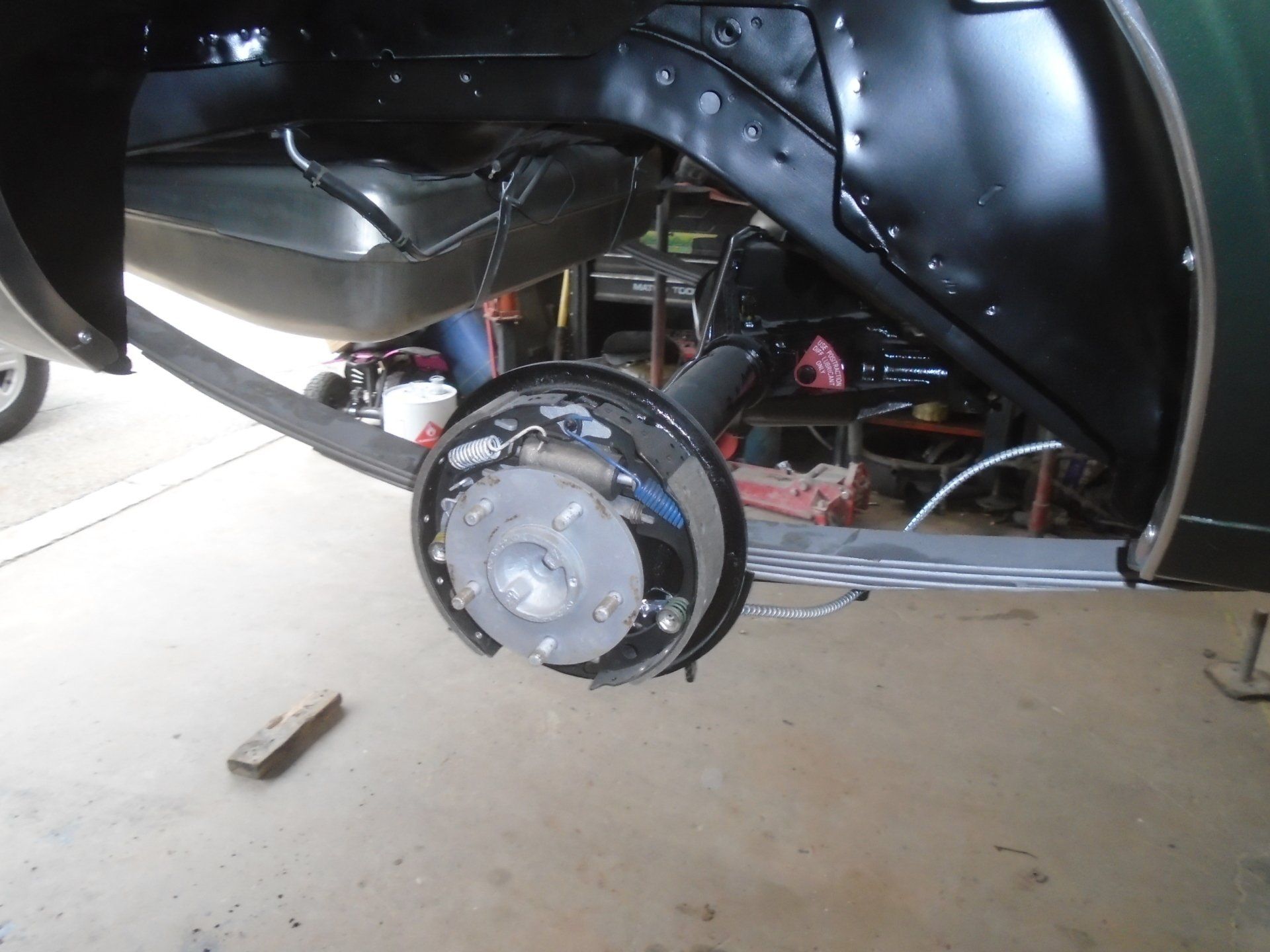 Car's rear wheel assembly, showing brake drum, axle, and leaf spring, inside a black wheel well.