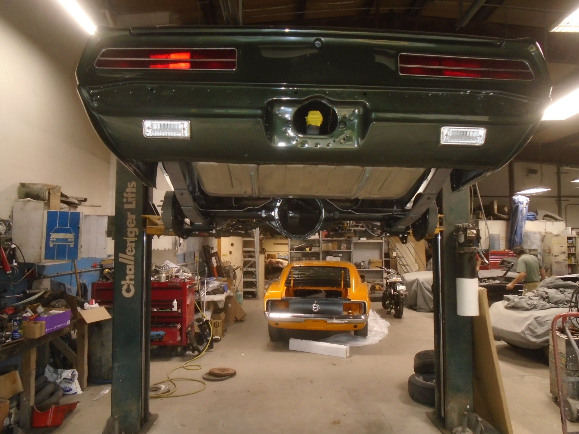 Dark green classic car on a lift in a garage, another yellow car in the background.