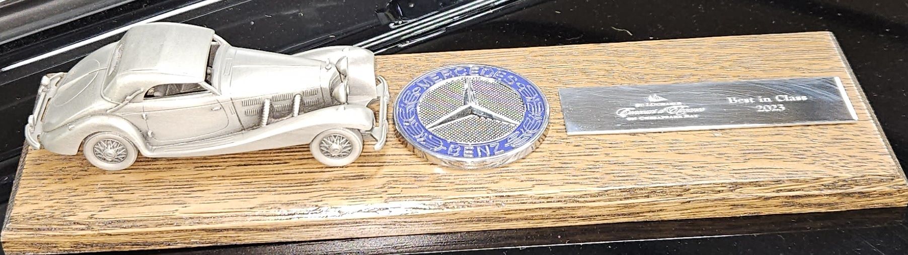 Miniature silver car, Mercedes logo, and plaque on a wooden base.