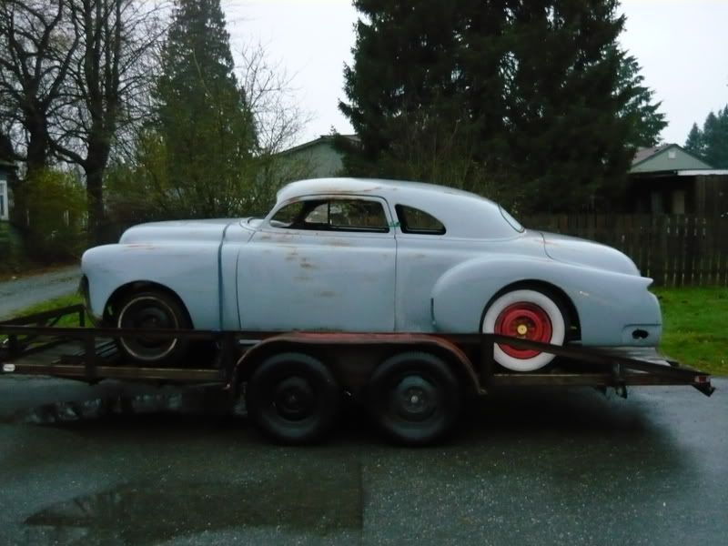 Classic light blue car on a trailer, with whitewall tires and red rims, parked on a wet street.