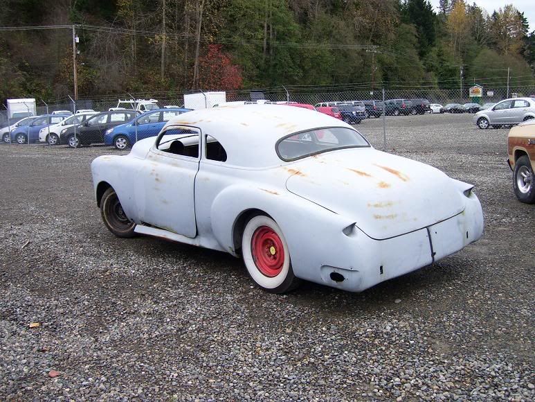 Grey classic car with white-wall tires parked on gravel. Red rims visible.
