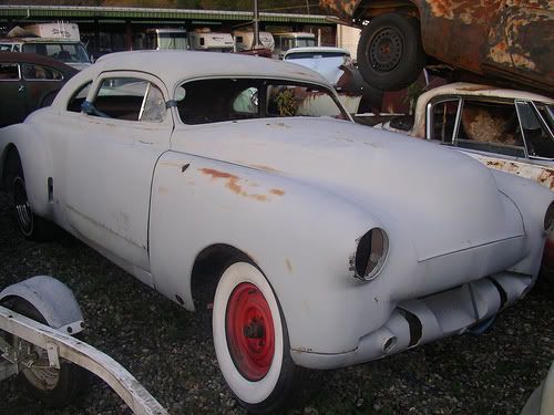 Grey classic car in a junkyard with red wheels and whitewall tires, rusting.