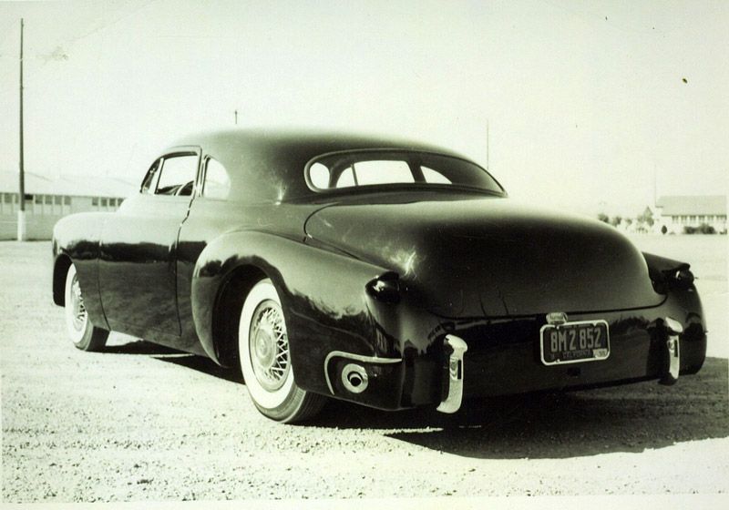 Black custom car, rear view, parked on a gravel lot, vintage style with white-wall tires.