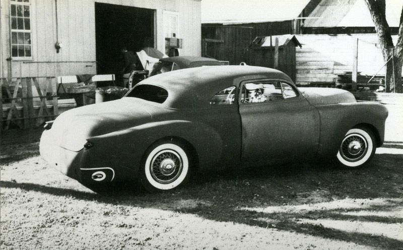 Custom car with chopped roof and white wall tires, parked near a wooden structure.