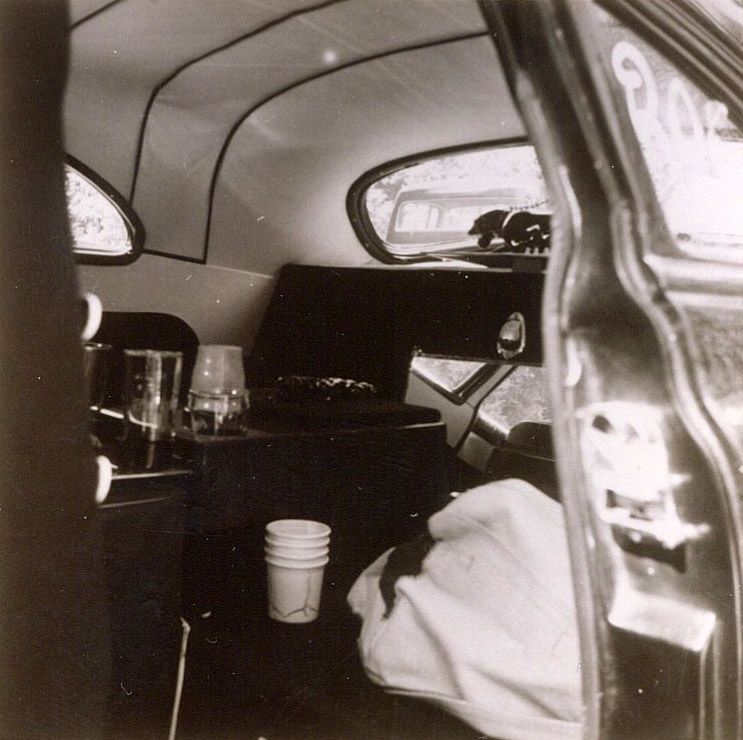 Interior of a vintage car with a dark interior, including seats, a beverage station, and a rear window.