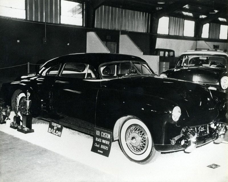 Black custom car, “Ed Cotton - Black Pearl”, displayed on a white platform, indoors.