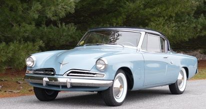 Light blue 1950s Studebaker coupe parked on a paved road, white-walled tires, black top, with trees in the background.
