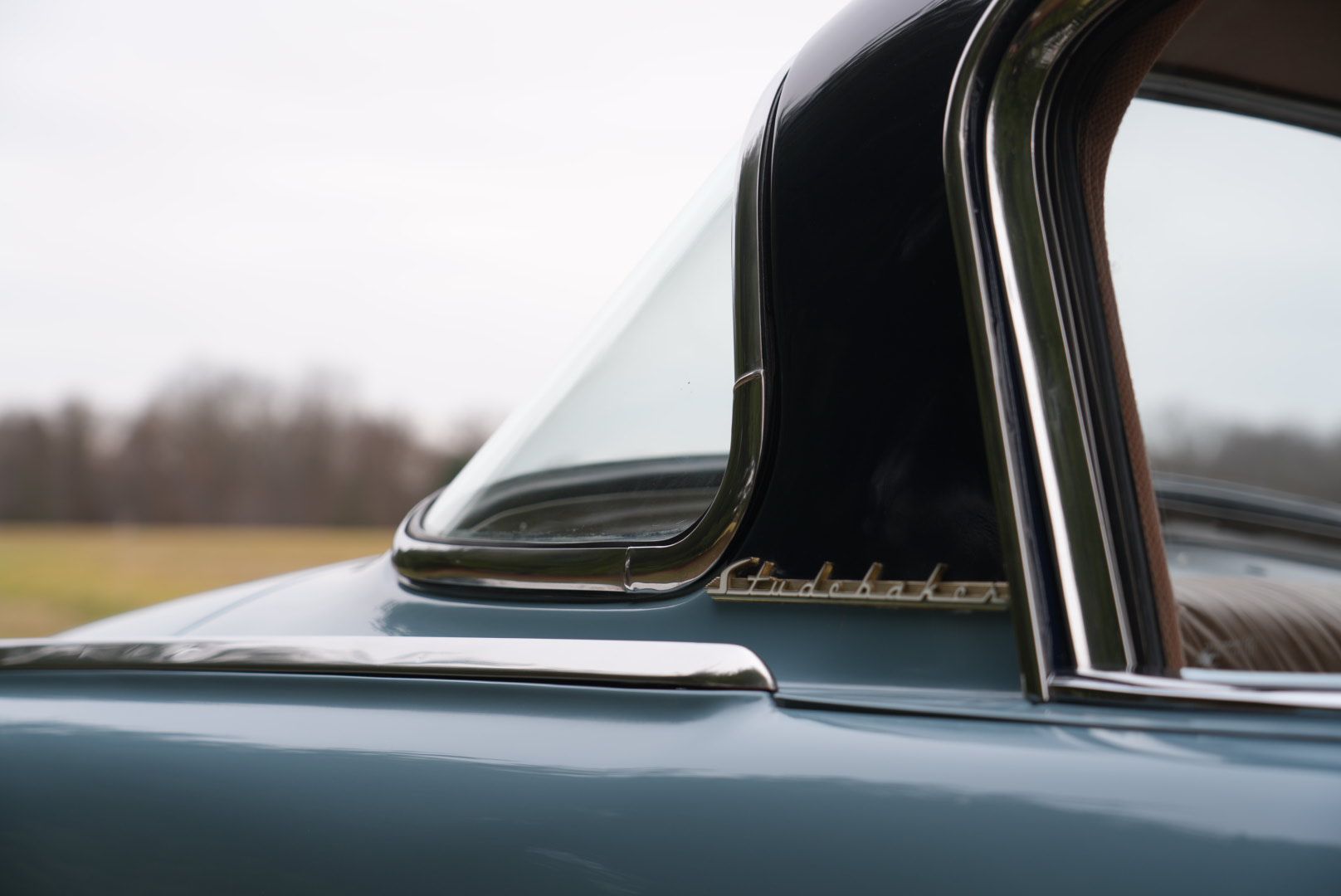 Blue Studebaker car with black roof and chrome trim; side view of the rear window and logo.