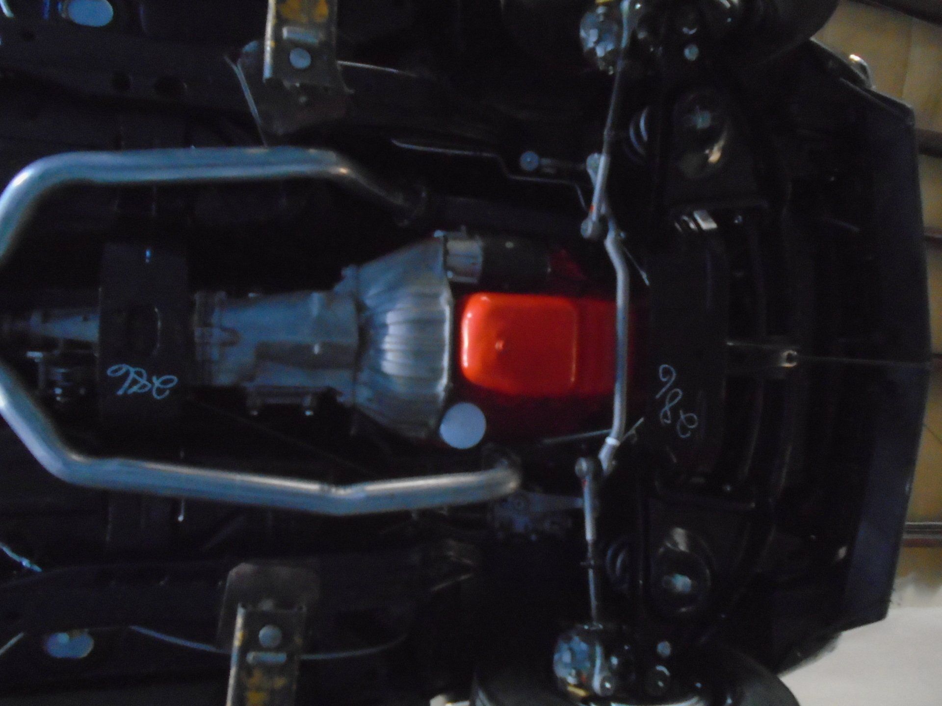 Underside view of a black classic car chassis, showing engine, transmission, and exhaust pipes.