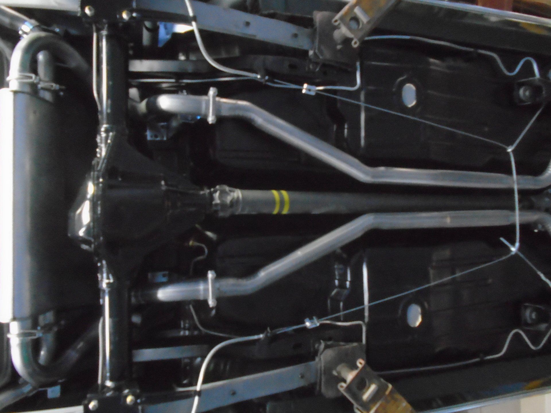 Underside of a black car with silver exhaust pipes, brake lines, and a yellow-striped driveshaft, supported on a lift.