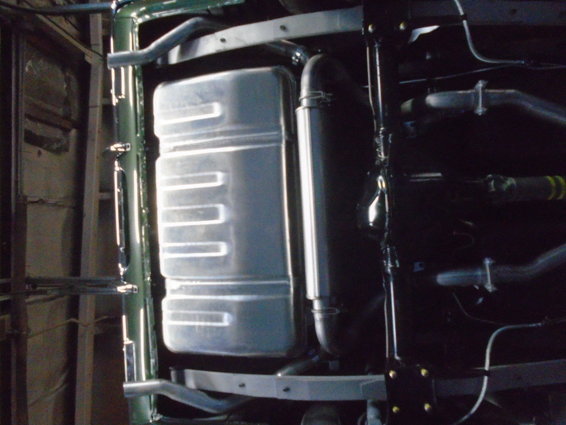 Undercarriage of a green vehicle showing the fuel tank, exhaust, and suspension components, mounted on a metal frame.