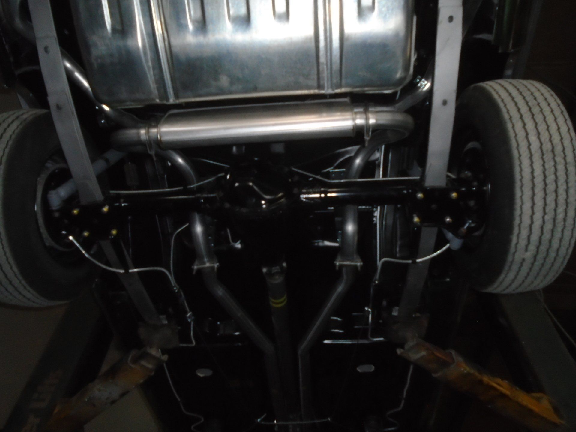 Undercarriage of a classic car, showing exhaust system, suspension, fuel tank, and tires. Black and silver components.