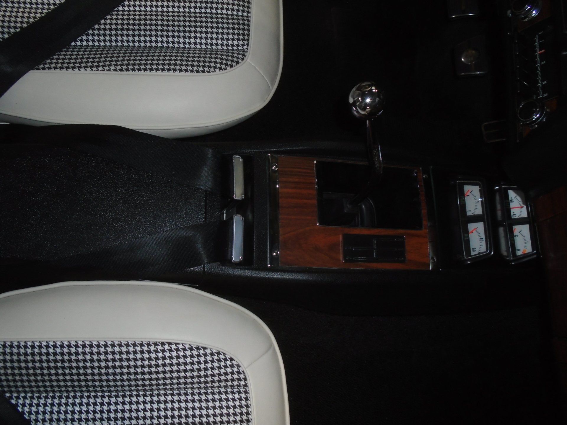 Close-up of a vintage car interior. Black and white patterned seats, wooden console, chrome shifter, and white trim.