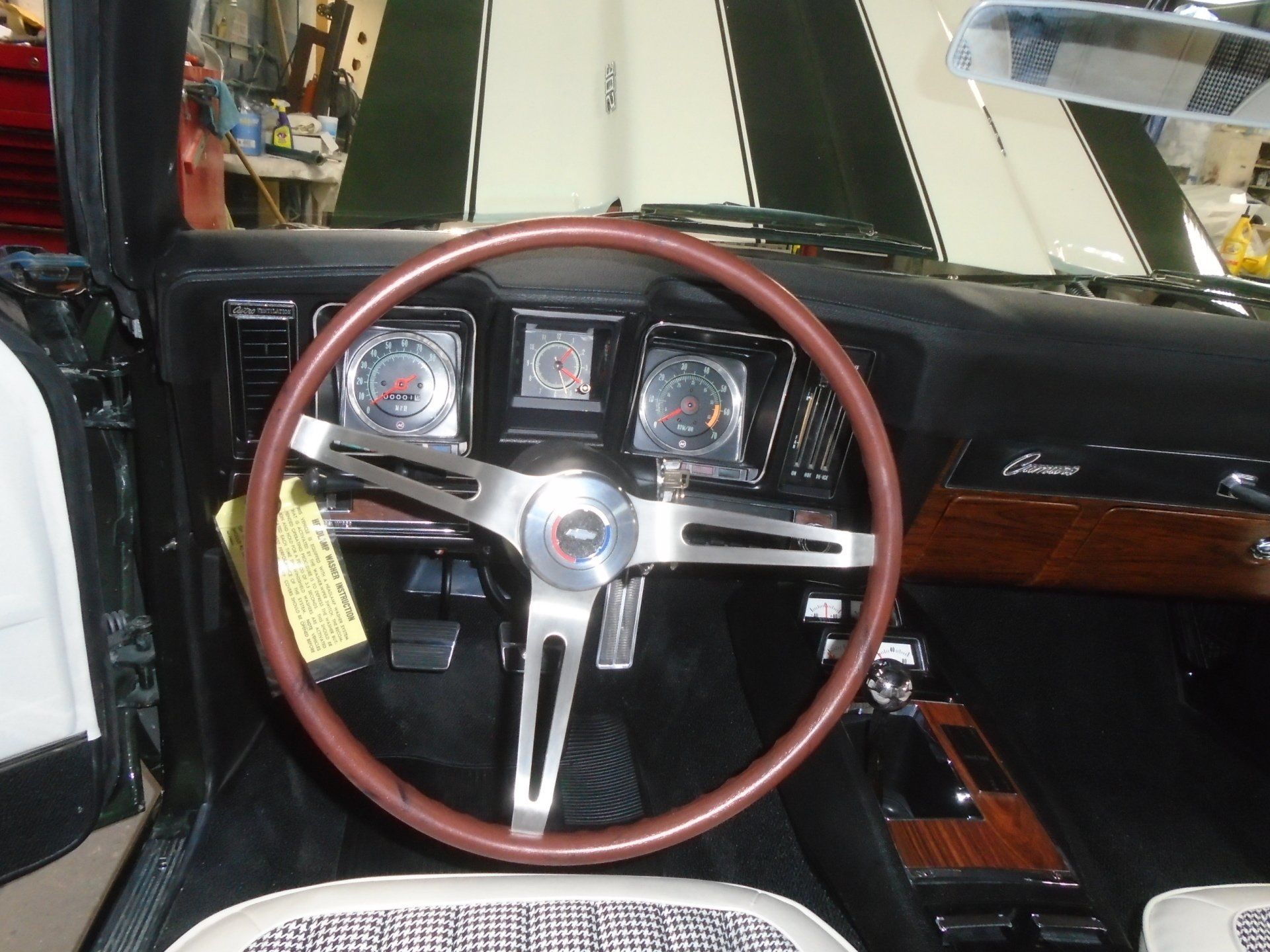 Interior of a classic black car with wood accents; steering wheel, gauges, and white and black patterned seat.