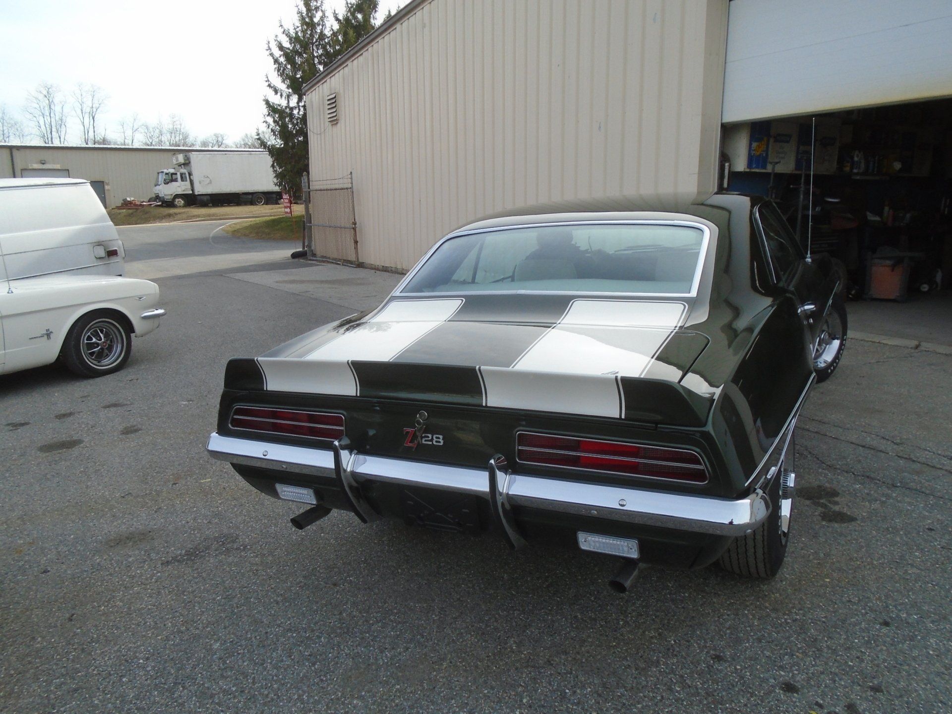 Black 1969 Camaro with white racing stripes in front of a garage. A white Mustang is to the left.