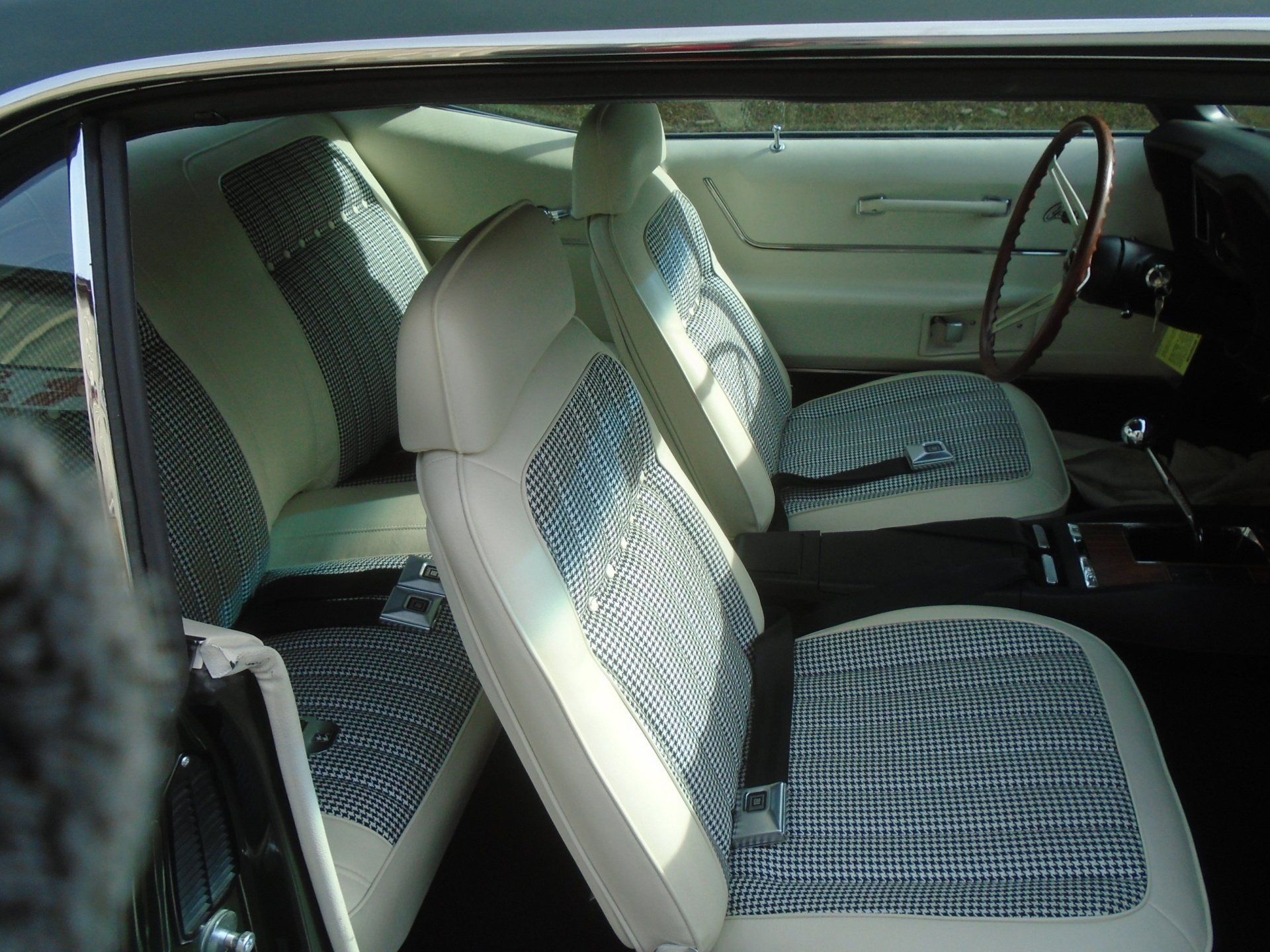 Interior view of a vintage car with cream and patterned seats, wood-grain steering wheel, and a manual gear stick.