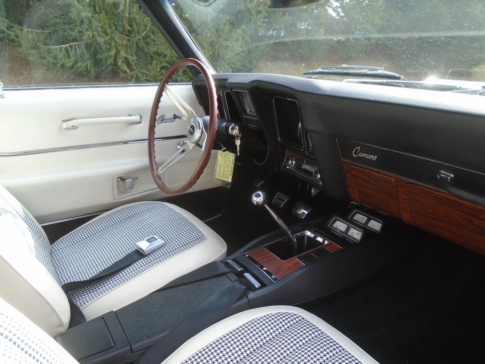 Interior view of a classic black and white Camaro. Features checkered seats, wooden accents, and a steering wheel.