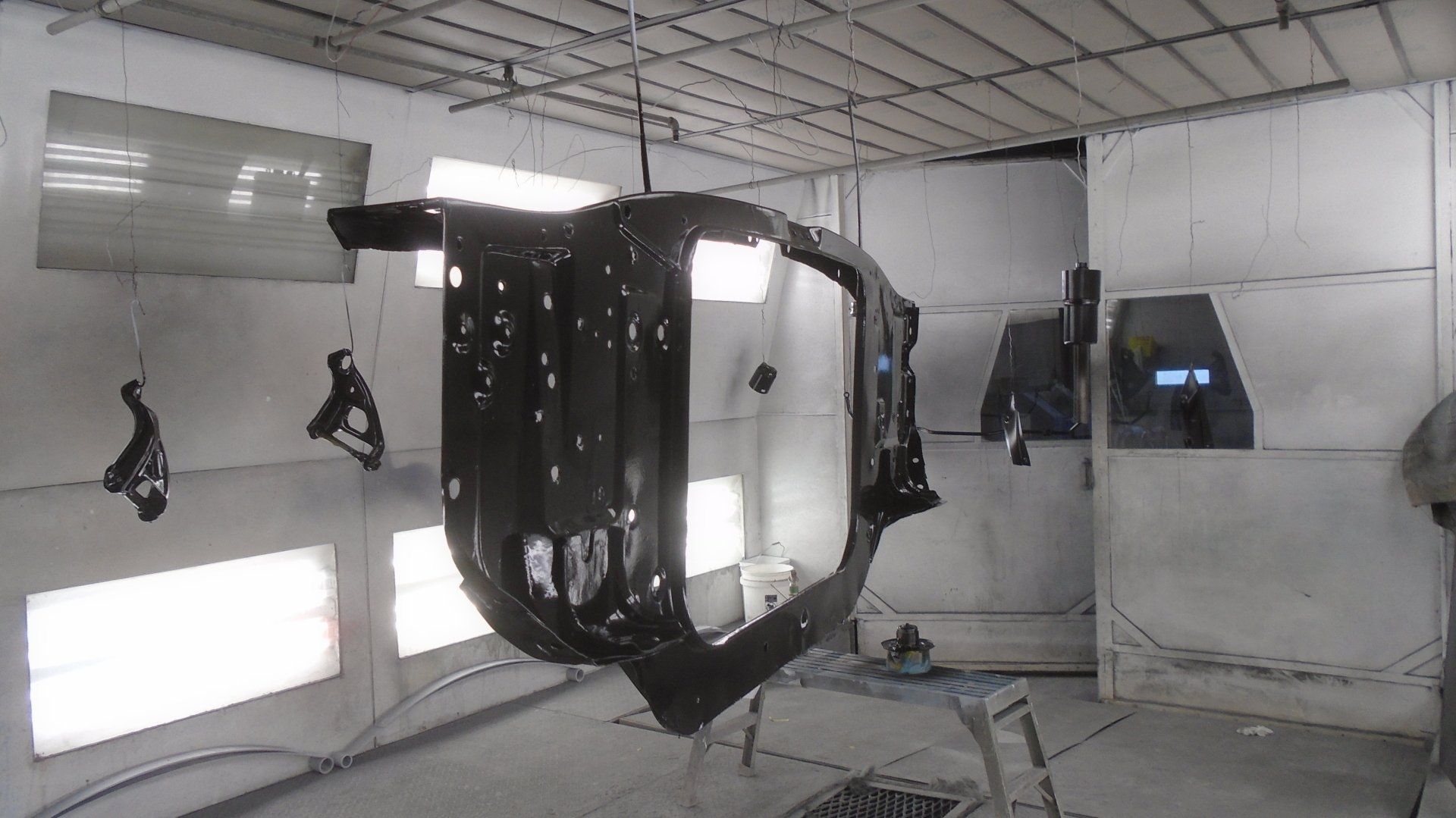 Car body part suspended in a spray booth, painted black. Other parts hang nearby.