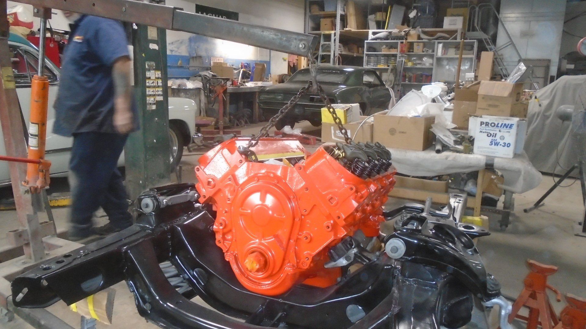 Bright orange car engine mounted on a black frame in a garage, a mechanic nearby.