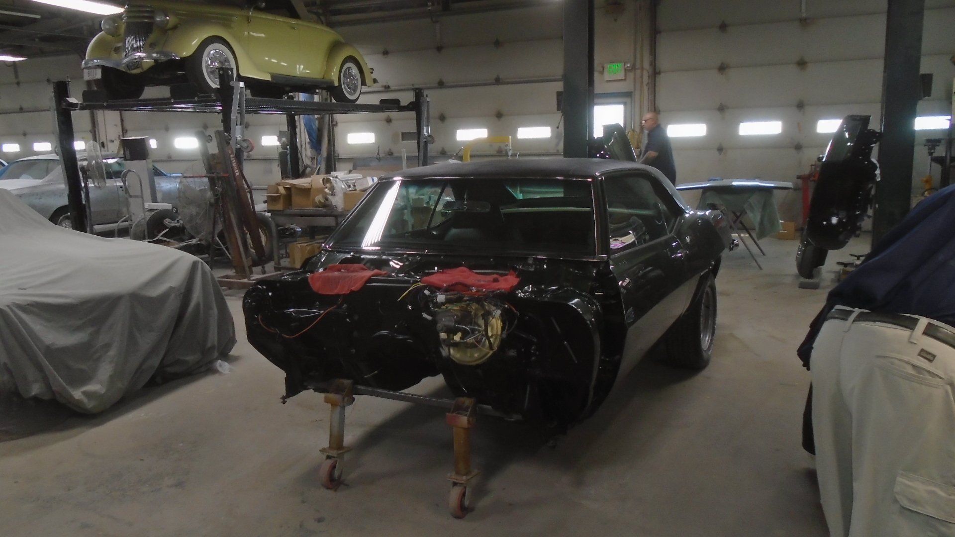 Black car body on stands in a garage; other cars, tools, and equipment are visible.