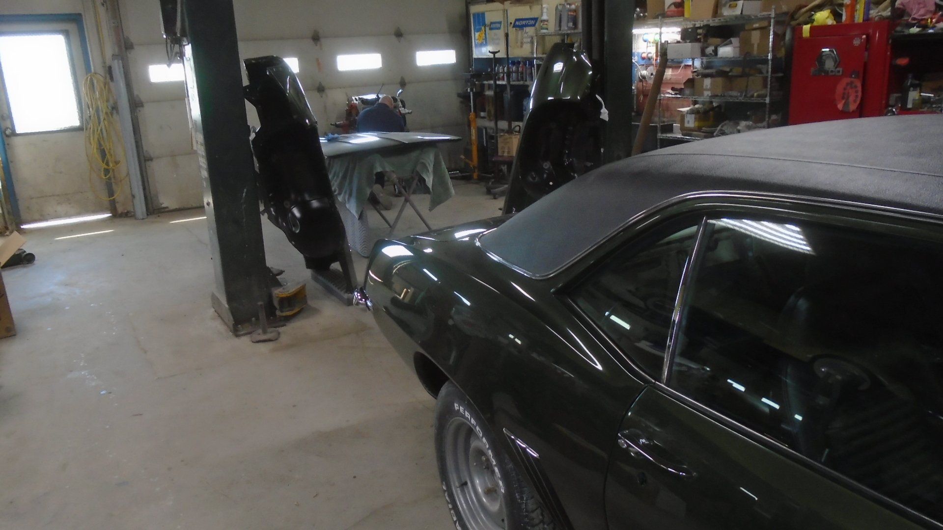 Dark green classic car in a workshop, near a car lift, with open doors and a covered table.