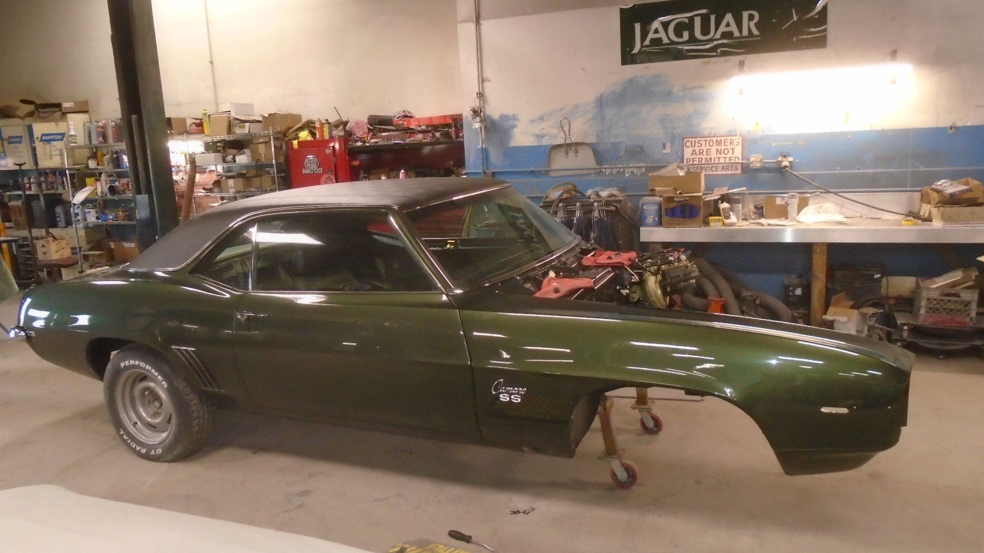 Green 1969 Chevrolet Camaro in a garage, missing front fender. Jaguar sign visible in the background.