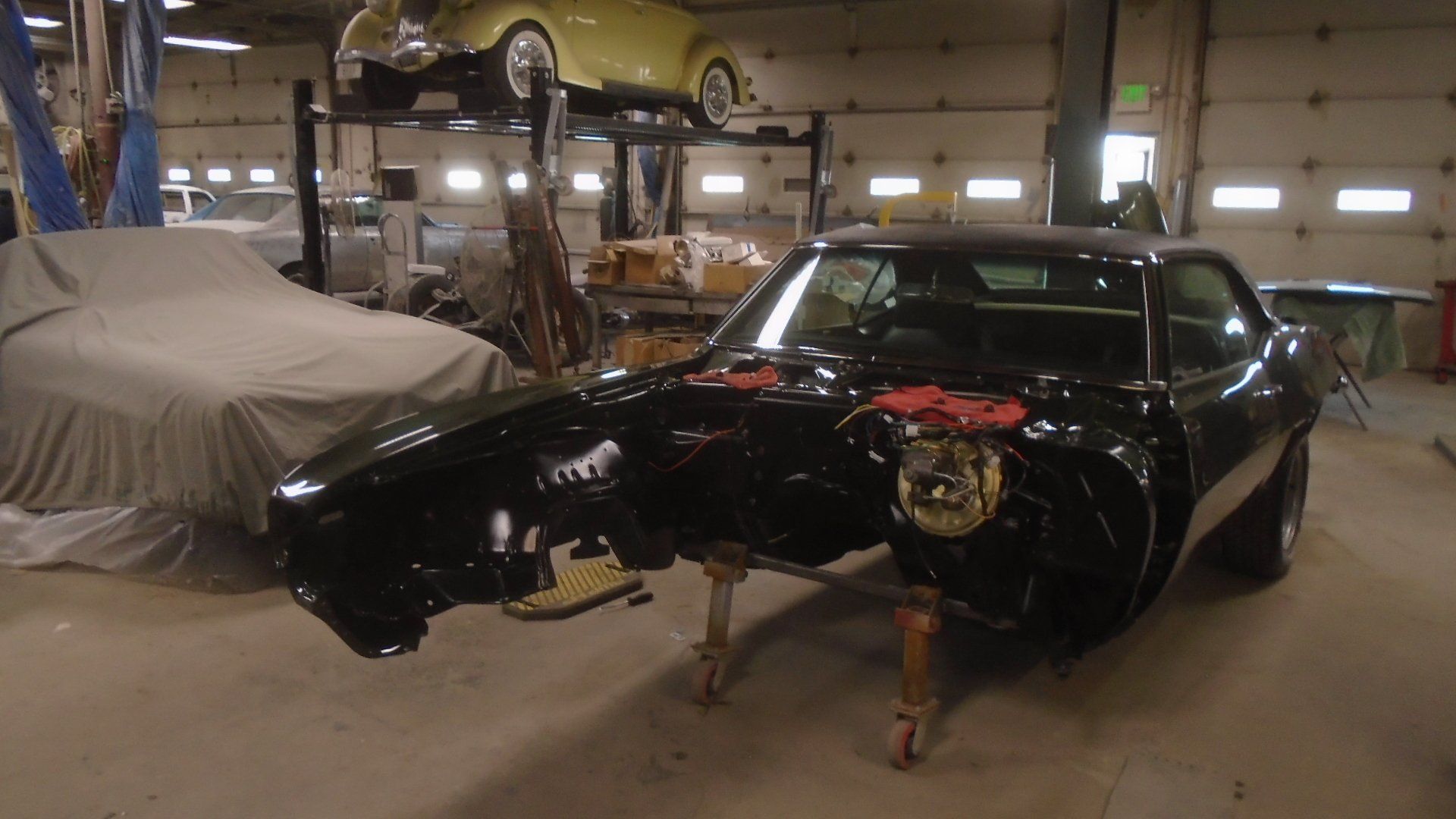 Black classic car in a workshop, partially disassembled. Another car covered, yellow car above.