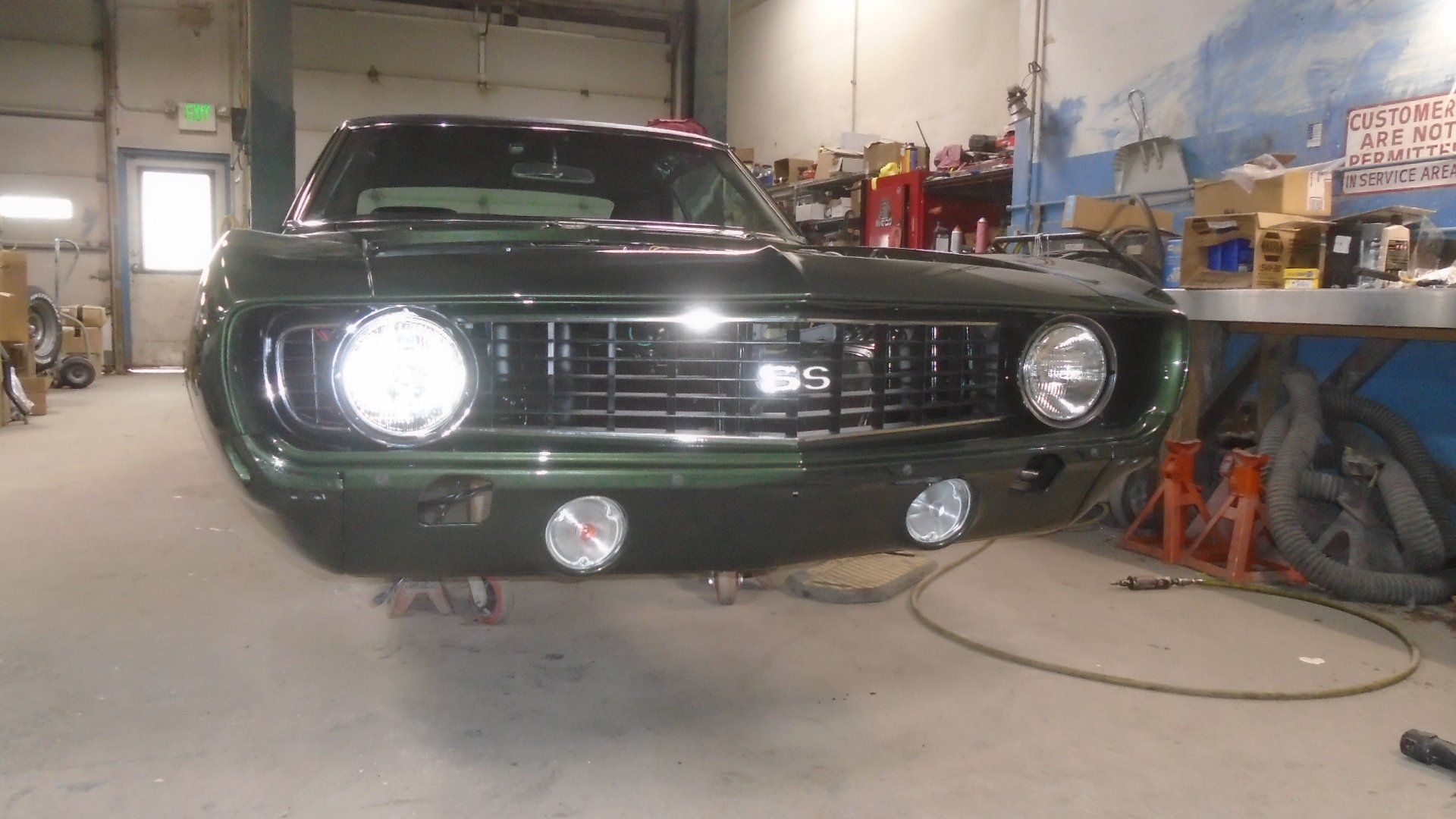 Green classic car with bright headlights and fog lights in a garage setting.