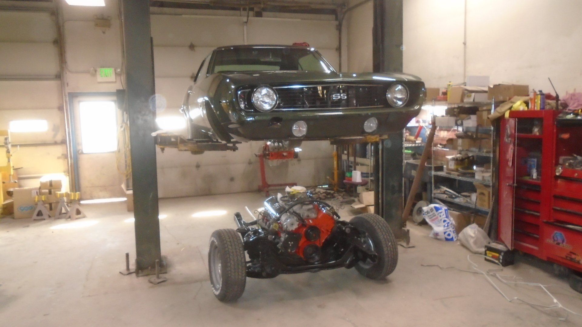 Dark green car body lifted over a chassis with an engine in a garage setting.