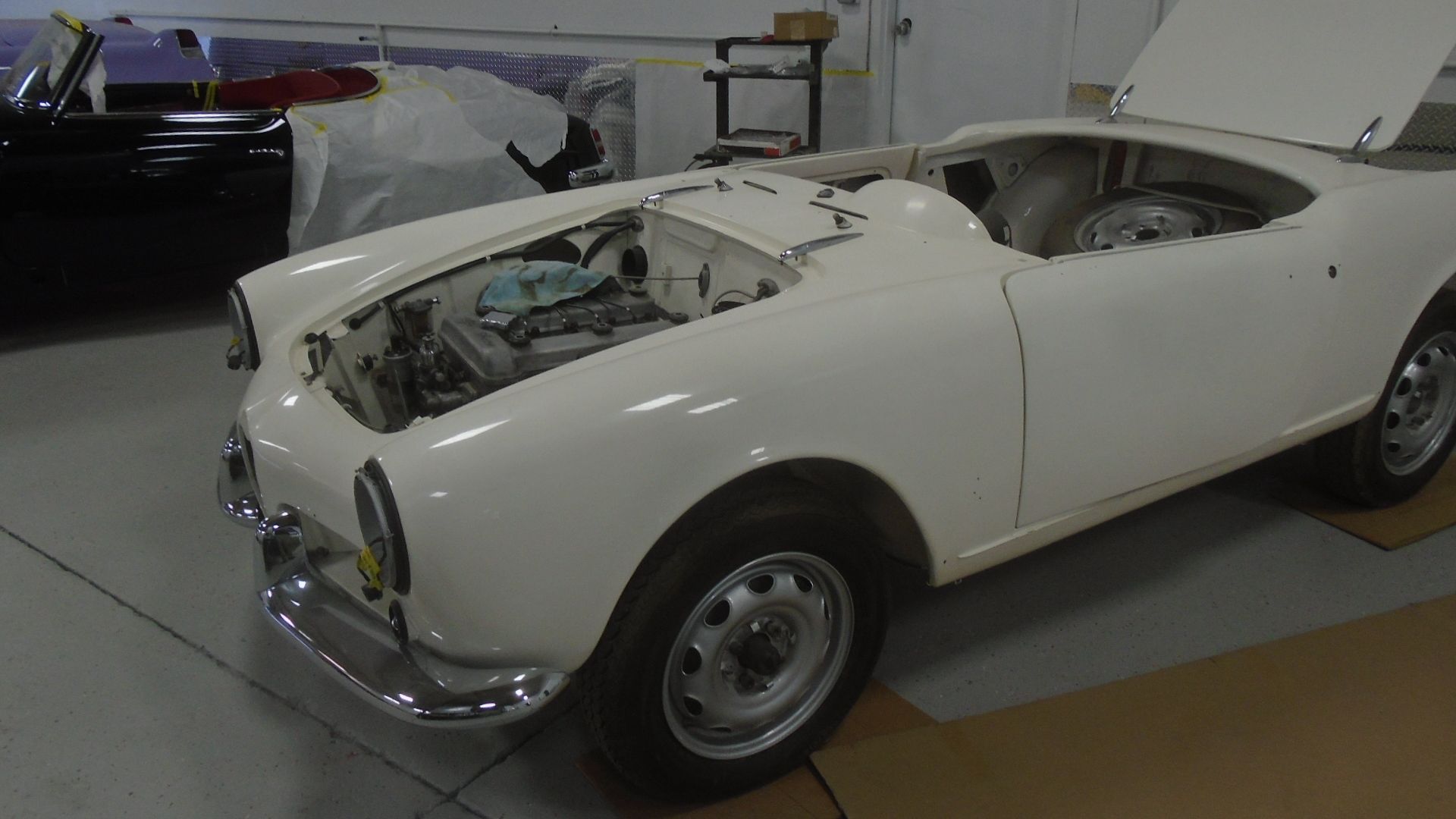 White classic car in a garage with hood and trunk open, showing the engine and spare tire.