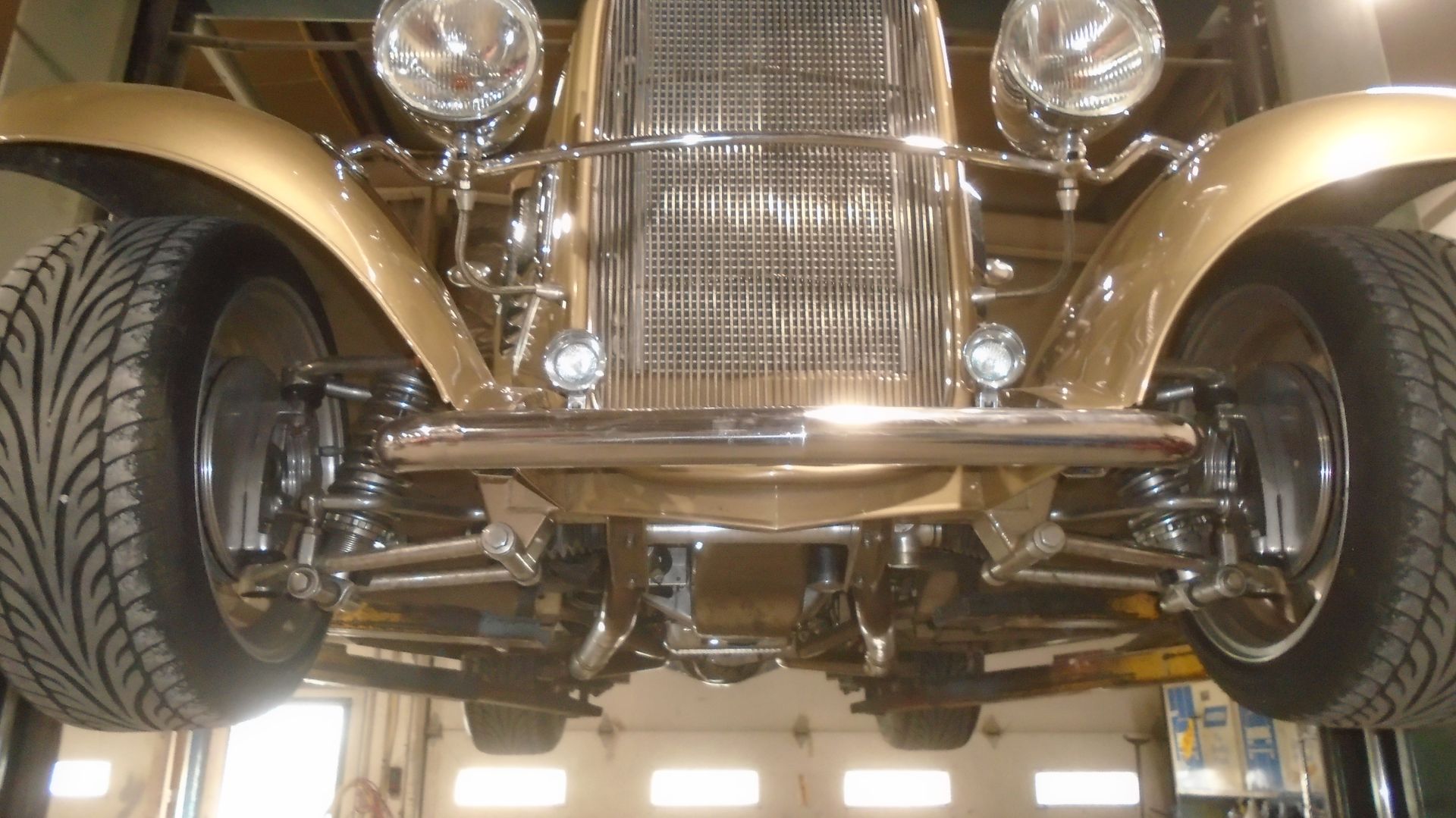Gold classic car lifted, showing undercarriage. Chrome details, large tires, and bright lights.
