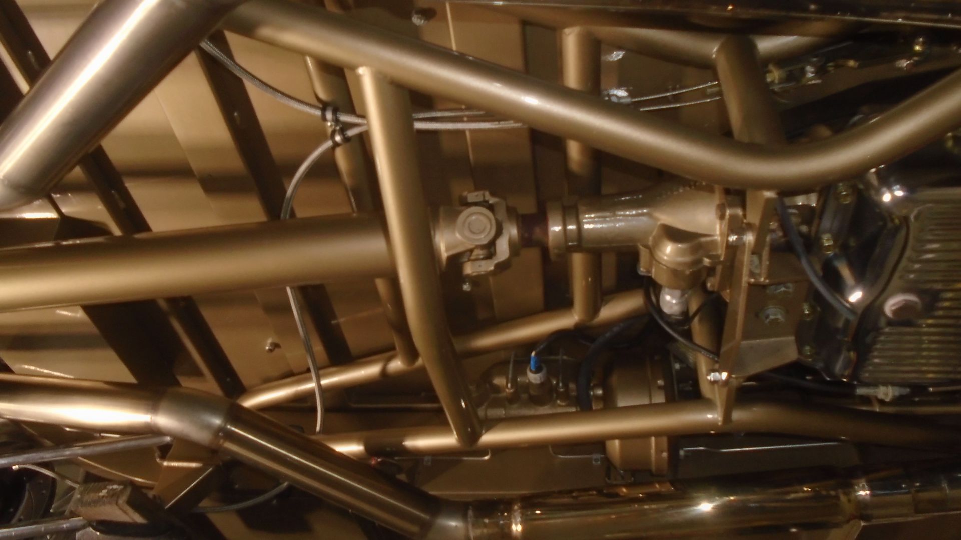 Undercarriage of a golden-colored car, featuring a driveshaft, various tubes, and supports.