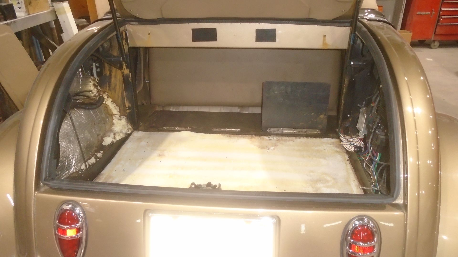 Open trunk of a gold classic car with interior exposed; gray metal floor, tan walls.