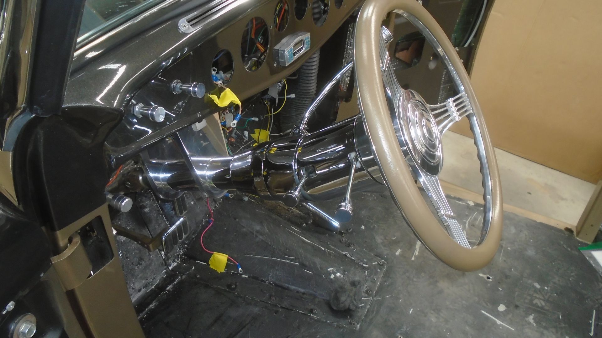 Interior of a vintage car with chrome steering wheel, dashboard, and gauges. Black and tan color scheme.