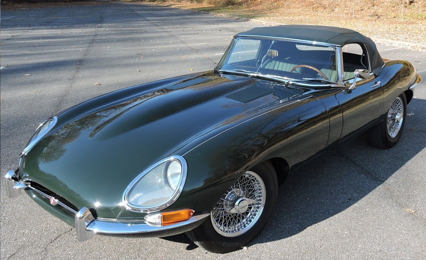 Green Jaguar E-Type convertible parked on a paved surface. Chrome accents and wire wheels.