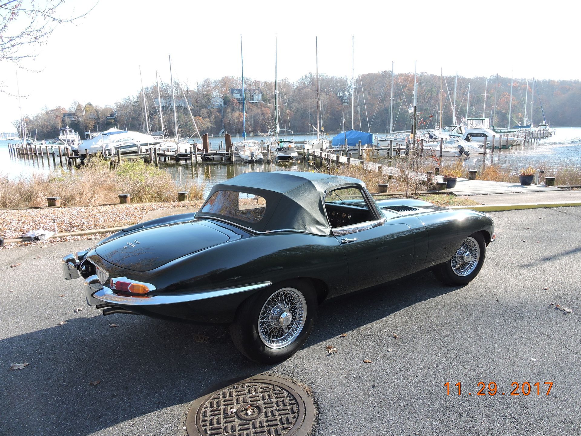 Green Jaguar E-Type convertible parked on asphalt by a marina. Black soft top is up. Date stamp is visible.